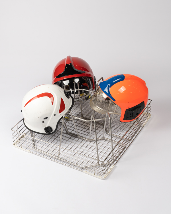Trois casques de pompier — blanc, rouge et orange — placés dans un support métallique pour SCBA et casques avec compartiments en fil métallique.