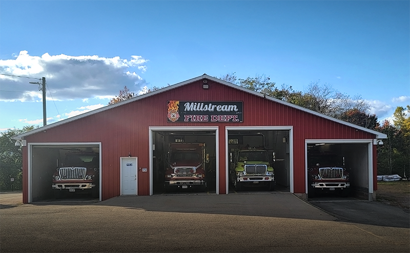 Red fire station building with three open bays showing fire trucks, sign reads Millstream Fire Dept.