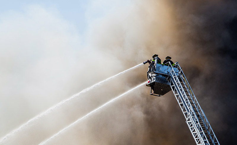 Feuerwehrleute auf einem Feuerwehrkran beim Löschen eines Brandes, wobei sie PAK und anderen giftigen Chemikalien ausgesetzt sind.